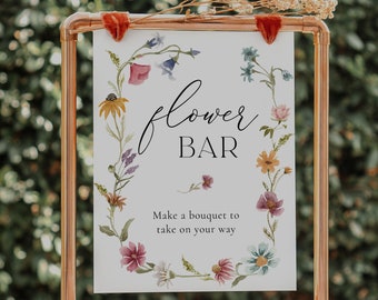 May include: A copper-colored metal frame holds a sign that reads "flower BAR" in elegant script. The sign is decorated with a floral wreath and the text "Make a bouquet to take on your way." Dried flowers rest on top.