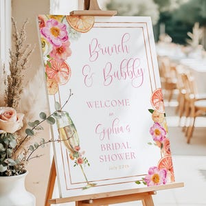May include: A white sign on a wooden easel with the text "Brunch & Bubbly" and "Welcome to Sophia's Bridal Shower July 20th". The sign is decorated with watercolor flowers and orange slices.