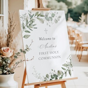 Könnte beinhalten: Ein weißes Schild auf einer hölzernen Staffelei mit dem Text "Welcome to Amelia's First Holy Communion". Das Schild ist mit grünen und grauen Aquarell-Eukalyptusblättern und einem Kreuz verziert. Eine Vase mit Blumen befindet sich im Vordergrund.