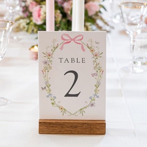 May include: A white card with a pink ribbon and floral wreath design. The card says "Table 2" in black text.