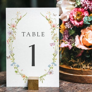 May include: A white card with a floral wreath design and the text "TABLE 1" printed on it. The card is sitting on a wooden table with a floral arrangement in the background.