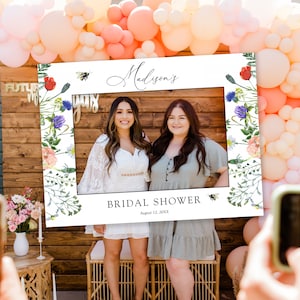 May include: White photo booth frame with a floral border and the text "Madison's Bridal Shower August 12, 20XX". Two women are posing in the frame.