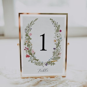 May include: A gold-framed table number card with a floral wreath design. The number "1" is displayed in black font. The text "Table" is written in cursive below the number.
