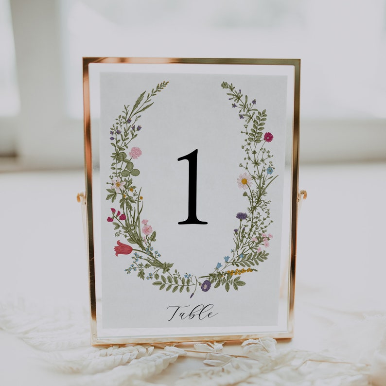 May include: A gold-framed table number card with a floral wreath design. The number "1" is displayed in black font. The text "Table" is written in cursive below the number.