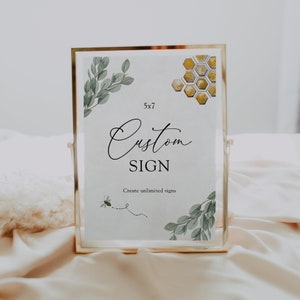 May include: A gold-framed sign with a white background and the text "Custom Sign" in black cursive font. The sign also features a bee illustration, green leaves, and a honeycomb pattern.