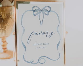 May include: A framed sign with a gold-colored frame. The sign has a white background with blue watercolor accents. The text "favors, please take a treat" is written in a delicate script font. A blue bow and ribbon design frames the text.