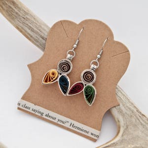 May include: A pair of dangle earrings featuring paper quilling in circular and teardrop shapes. The colors include dark brown, yellow, teal, red, and green. The earrings are attached to a tan card with the text "is class saying about you?" Hermione won.