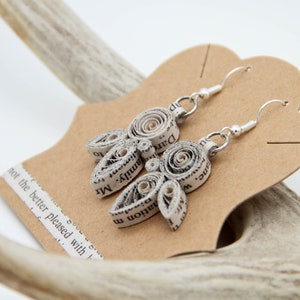 May include: A pair of dangle earrings crafted from coiled strips of printed book pages. Each earring features a central circular coil surrounded by three smaller teardrop-shaped coils, all attached to silver-toned metal hooks for pierced ears.