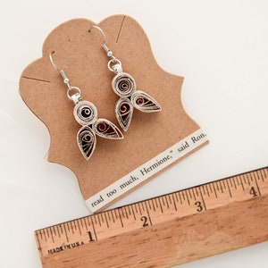 May include: A pair of silver-toned quilled paper earrings featuring a spiral and teardrop design with dark red centers. The earrings are attached to a tan card with the text "read too much, Hermione," said Ron," next to a wooden ruler showing 5 inches.
