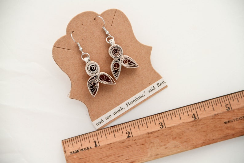 May include: A pair of silver-toned quilled paper earrings featuring a spiral and teardrop design with dark red centers. The earrings are attached to a tan card with the text "read too much, Hermione," said Ron," next to a wooden ruler showing 5 inches.