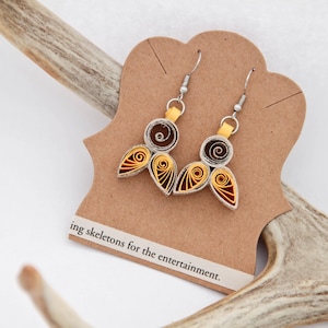 May include: A pair of dangle earrings made from coiled paper strips in shades of dark brown and golden yellow. Each earring features a circular top and two teardrop-shaped bottom pieces attached to silver-toned metal hooks on a brown card.