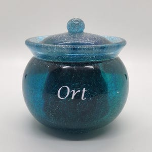May include: A round, lidded jar with a glittery teal and dark blue ombre design. The jar has a matching lid with a small knob. The word "Ort" is written in white on the front of the jar. The jar is likely used for storage.