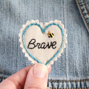 Puede incluir: Un parche de tela blanco en forma de corazón con costuras azules alrededor de los bordes. La palabra "Brave" está bordada en hilo negro en el parche. Una pequeña abeja bordada amarilla y negra está sobre la palabra.
