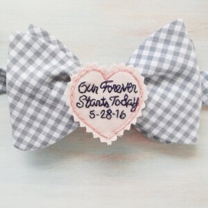 May include: A gray and white gingham bow tie with a pink heart-shaped patch that says "Our Forever Starts Today 5-28-16" in black embroidery.