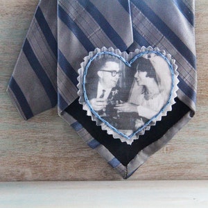 Puede incluir: Una corbata a rayas grises y azules con un parche de tela en forma de corazón cosido. El parche presenta una fotografía en blanco y negro de una pareja.