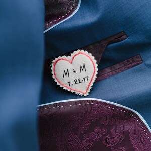 Könnte beinhalten: Ein weißer herzförmiger Stoff-Patch mit roten Nähten, auf einen blauen Stoffhintergrund genäht. Der Patch trägt den Text "M + M 7.22.17", der in schwarzem Garn gestickt ist.