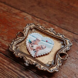 May include: A heart-shaped fabric patch with a photo of a couple on a beach, embroidered with the date "12.2.23", displayed in a gold-toned ornate tray.