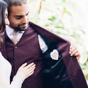 Può includere: Una coppia in un contesto di matrimonio. Lo sposo indossa una giacca e un gilet color borgogna, e tiene la giacca aperta per rivelare una toppa di stoffa a forma di cuore cucita all'interno. La toppa ha le parole "You Are My Everything" scritte su di essa.
