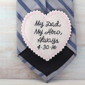 Könnte beinhalten: Ein weißer herzförmiger Stoff-Patch mit rosa Nähten an den Rändern. Der Patch ist mit schwarzer Stickerei versehen, die "My Dad, My Hero, Always 4-30-76" lautet.