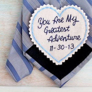 Könnte beinhalten: Ein weißer herzförmiger Stoff-Patch mit blauen Nähten an den Rändern. Der Patch hat den Text "You are my greatest adventure 11-30-13" in schwarzem Garn gestickt.