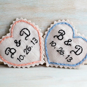 Könnte beinhalten: Zwei herzförmige Stoffschmuckstücke mit gestickten Initialen und einem Datum. Das linke Ornament ist rosa mit den Initialen "B & D" und dem Datum "10.26.13". Das rechte Ornament ist blau mit den Initialen "B & D" und dem Datum "10.26.13".