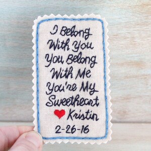 Könnte beinhalten: Ein weißes Stoffquadrat mit blauen Nähten an den Rändern. Der Stoff ist mit schwarzer Stickerei versehen, die "I belong with you, you belong with me, you're my sweetheart Kristin 2-26-16" lautet. Ein kleines rotes Herz ist über dem Namen gestickt.