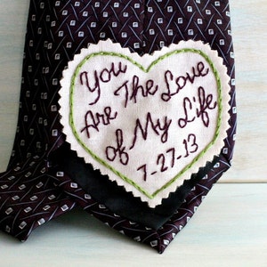 Könnte beinhalten: Ein weißer herzförmiger Stoff-Patch mit grüner Steppnaht um die Ränder. Der Patch ist mit dem Text "You are the love of my life" und dem Datum "7-27-13" bestickt. Der Patch ist an einer dunkelbraunen gemusterten Krawatte befestigt.