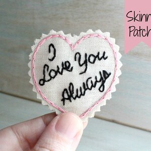 Könnte beinhalten: Ein weißer herzförmiger Stoff-Patch mit rosa Nähten an den Rändern. Der Patch ist mit den Worten "I love you always" in schwarzem Faden bestickt.