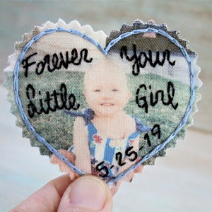 May include: A heart-shaped fabric patch with a photo of a baby girl and the text "Forever my little girl 6-25-19" embroidered in black thread. The heart is outlined with blue thread.