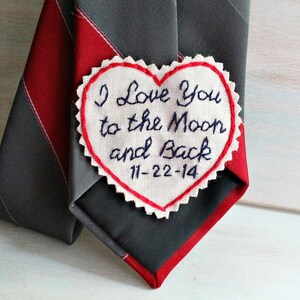 Könnte beinhalten: Ein weißer herzförmiger Stoff-Patch mit roten Nähten an den Rändern. Der Patch ist mit blauer Stickerei versehen, die "I love you to the moon and back 11-22-14" lautet. Der Patch ist an einer grau-rot gestreiften Krawatte befestigt.