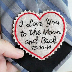 Könnte beinhalten: Ein weißer herzförmiger Stoff-Patch mit roten Nähten an den Rändern. Der Patch ist mit schwarzer Stickerei versehen, die "I love you to the moon and back 25-10-14" lautet.