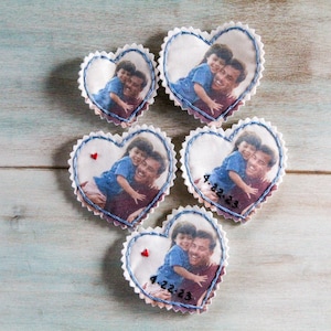 May include: Five white fabric heart-shaped ornaments with blue stitching around the edges. Each heart features a photo of a father and child, with the date "4.22.23" written on two of the hearts.