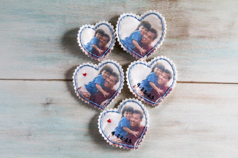 May include: Five white fabric heart-shaped ornaments with blue stitching around the edges. Each heart features a photo of a father and child, with the date "4.22.23" written on two of the hearts.