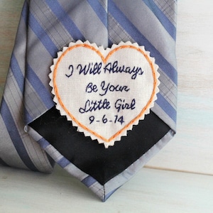 Könnte beinhalten: Ein weißer herzförmiger Stoff-Patch mit dem Text "I Will Always Be Your Little Girl 9-6-14", der in schwarzem Garn gestickt ist. Der Patch ist an einer grau-blauen gestreiften Krawatte befestigt.