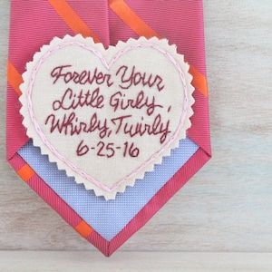 Könnte beinhalten: Ein rosafarbenes herzförmiges Stoffstück mit dem Text "Forever Your Little Girly, Whirly, Twirly" und dem Datum "6-25-16", das in kastanienbraunem Garn gestickt ist. Das Stück ist an einer rosa-orangefarbenen gestreiften Krawatte befestigt.
