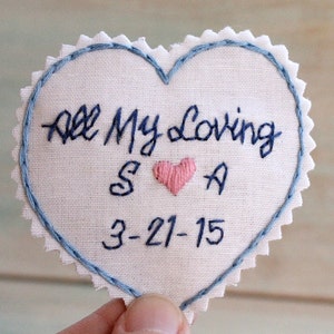 Può includere: Un cerotto di stoffa bianco a forma di cuore con cuciture blu intorno ai bordi. Il cerotto è ricamato con il testo "All My Loving" e le iniziali "S" e "A" con un cuore rosa tra loro. La data "3-21-15" è ricamata sotto le iniziali.