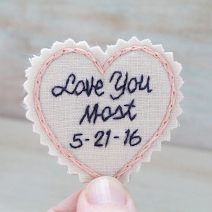 Könnte beinhalten: Ein rosa herzförmiger Stoff-Patch mit den Worten "Love You Most" und dem Datum "5-21-16", das in schwarzem Garn gestickt ist.