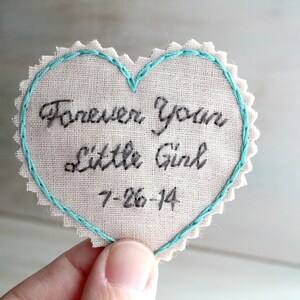 Könnte beinhalten: Ein herzförmiger Stoff-Patch mit einem hellblauen, gestickten Rand. Der Patch ist aus beigefarbenem Stoff und hat den Text "Forever Your Little Girl" und das Datum "7-26-14" in Schwarz gestickt.