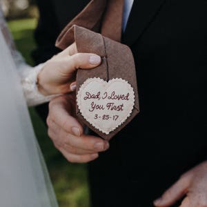 Könnte beinhalten: Eine braune Krawatte mit einem herzförmigen Anhänger, auf dem steht "Dad, I loved you first 3-25-17".