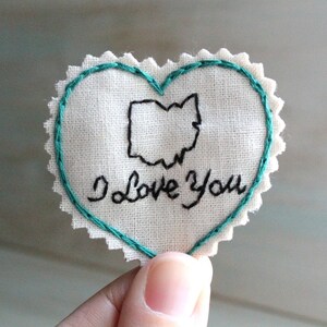 Könnte beinhalten: Ein weißer herzförmiger Stoff-Patch mit einem türkisfarbenen, gestickten Rand. Der Patch zeigt eine schwarze, gestickte Umrandung des Bundesstaates Ohio und den Text "Ich liebe dich".