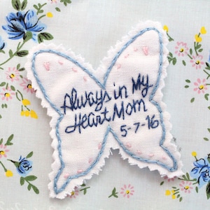 Puede incluir: Un parche de tela blanco con forma de mariposa con bordes bordados en azul. El parche tiene el texto "Always in my Heart Mom 5-7-16" bordado en hilo azul.