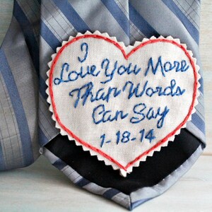 Könnte beinhalten: Ein weißer herzförmiger Stoff-Patch mit roten Nähten an den Rändern. Der Patch ist mit blauer Stickerei in kursiver Schrift mit dem Text "I Love You More Than Words Can Say 1-18-14" verziert. Der Patch ist an einer grau-blauen gestreiften Krawatte befestigt.
