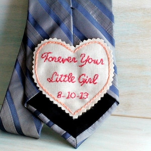 Könnte beinhalten: Eine graue und blaue gestreifte Krawatte mit einem weißen herzförmigen Patch, der darauf genäht ist. Der Patch ist in Pink bestickt und trägt die Aufschrift "Forever Your Little Girl 8-10-13".