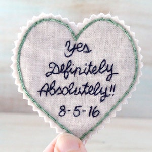 Könnte beinhalten: Ein weißer herzförmiger Stoff-Patch mit grüner Naht um die Ränder. Der Patch ist mit dem Text "Yes Definitely Absolutely!! 8-5-16" bestickt.