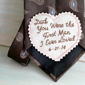 May include: A brown paisley tie with a white heart-shaped patch sewn on. The patch has pink stitching around the edges and brown embroidery that reads "Dad, you were the first man I ever loved 6-21-14".