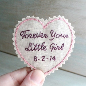 Könnte beinhalten: Ein rosa herzförmiger Stoff-Patch mit den Worten "Forever Your Little Girl" und dem Datum "8-2-14", das in lila Farbe gestickt ist.
