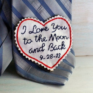 Könnte beinhalten: Ein weißer herzförmiger Stoff-Patch mit roten Nähten an den Rändern. Der Patch hat den Text "I love you to the moon and back" in schwarzem Garn gestickt. Das Datum "9-28-13" ist ebenfalls in schwarzem Garn unter dem Text gestickt.