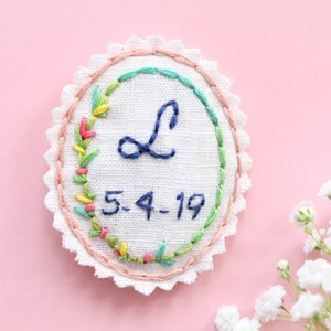 Peut inclure: Un patch brodé circulaire blanc avec une lettre L bleue, une couronne de fleurs et la date 5-4-19.