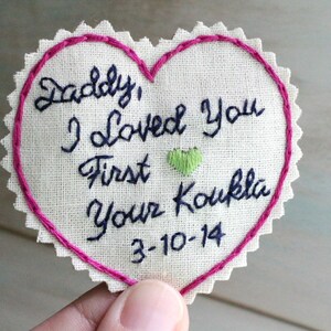 Könnte beinhalten: Ein herzförmiger Stoff-Patch mit rosa Nähten an den Rändern. Der Patch hat eine handschriftliche Botschaft in blauer Farbe, die lautet "Daddy, I loved you first Your Koukla 3-10-14".