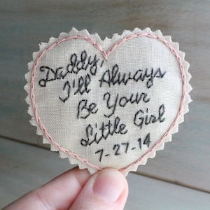Könnte beinhalten: Ein herzförmiges Stück Stoff mit rosa Nähten an den Rändern. Der Stoff ist mit schwarzem Garn bestickt mit den Worten "Daddy, I'll always be your little girl 7-27-14".